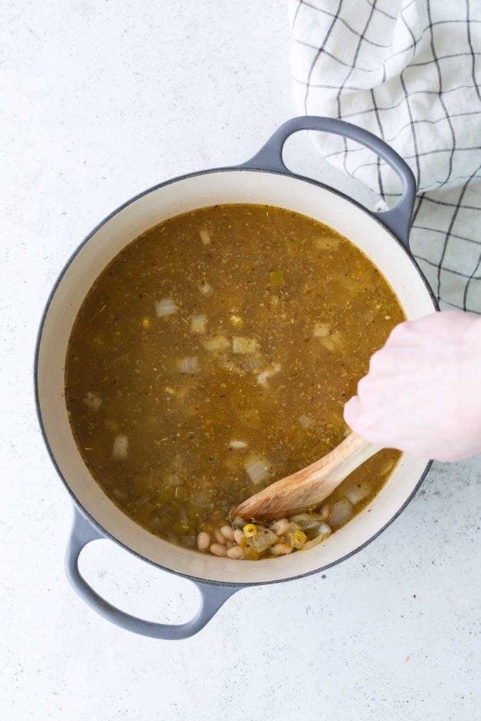 A hand stirs soup with beans and chopped vegetables in a large pot on a white surface, next to a checkered cloth.