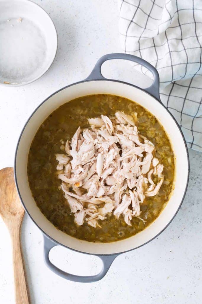 A pot of soup with shredded chicken on top, next to a wooden spoon and a checkered kitchen towel on a white surface.