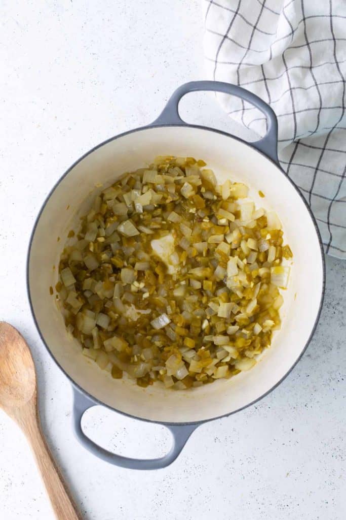 Chopped onions and green chilies sautéing in a large white pot, with a wooden spoon and a checkered towel nearby.