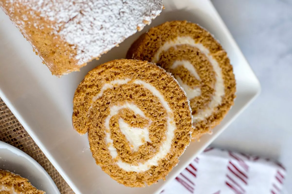 Two slices of pumpkin roll cake with cream cheese filling sit on a white plate, with a dusting of powdered sugar on top.