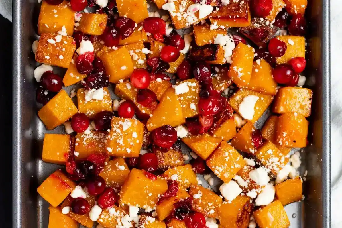 Roasted butternut squash cubes with cranberries and crumbled cheese on a baking sheet.