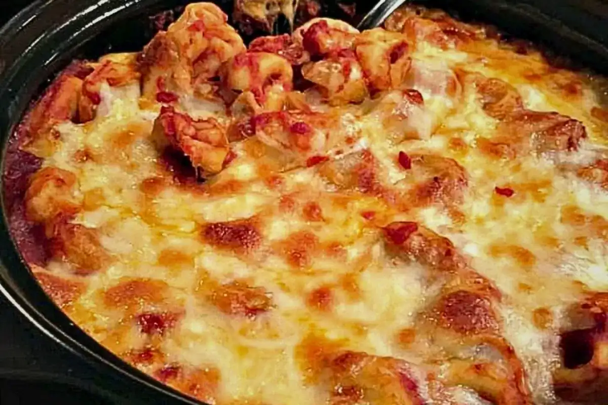 A close-up of a baked dish topped with melted cheese, tomato sauce, and pieces of pasta or meat in a black casserole dish.
