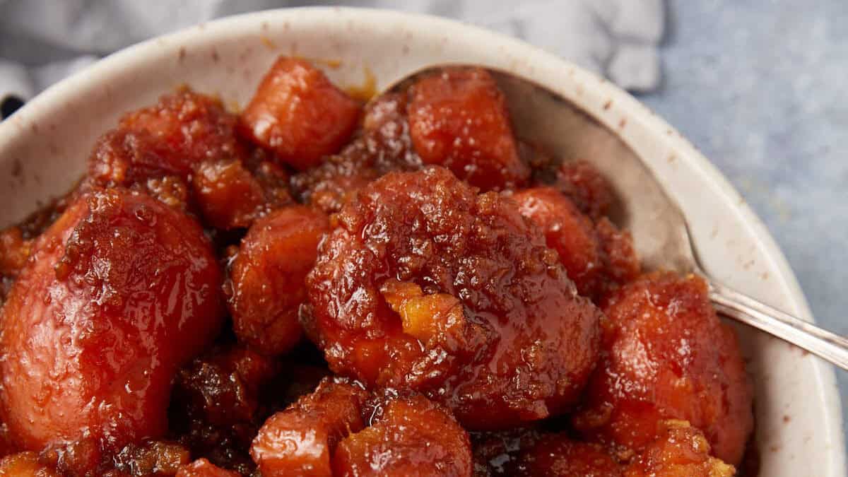 A bowl of candied yams in syrup with a spoon, showing glazed orange chunks and whole pieces.