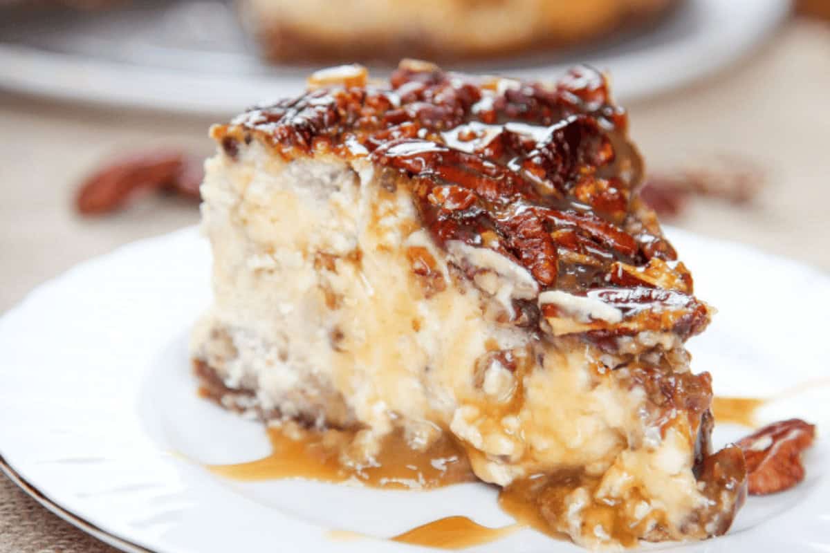 A slice of pecan-topped cheesecake with caramel sauce drizzled over it on a white plate.