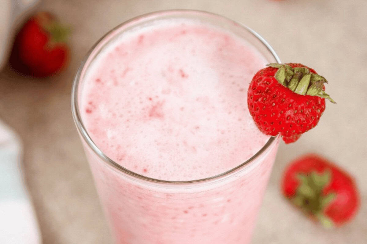 A glass of pink strawberry smoothie topped with a whole strawberry on the rim, with another strawberry out of focus in the background.