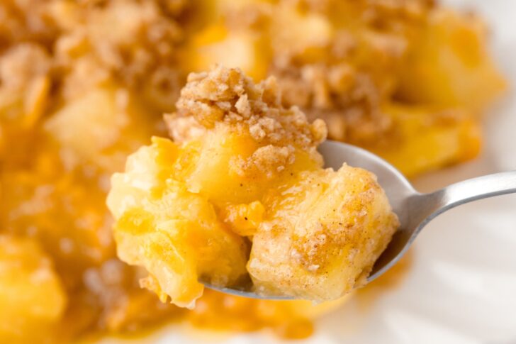 A close-up of a spoonful of pineapple casserole topped with a crumbly brown sugar and cracker topping, with more casserole visible in the background.