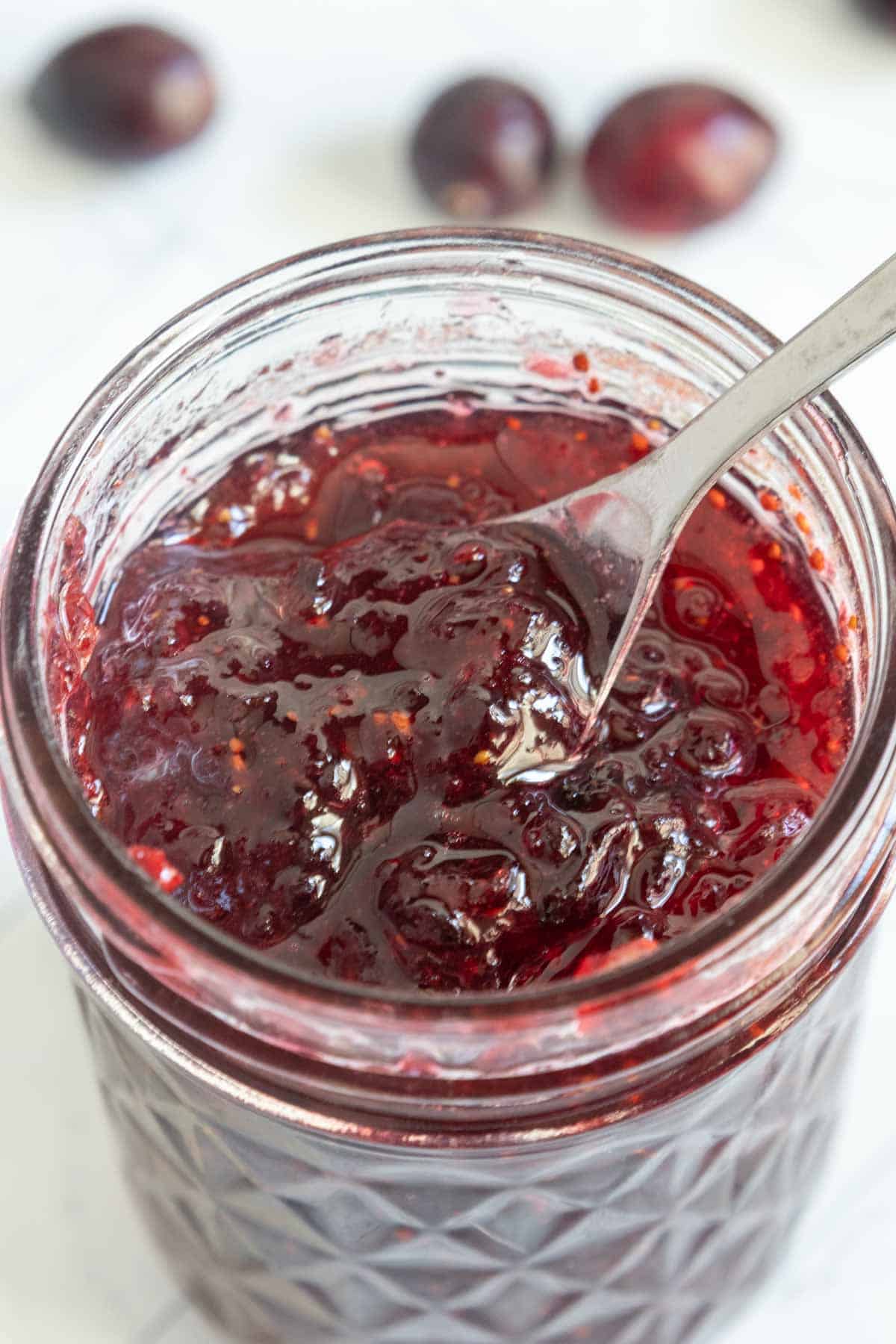 Open jar of dark red plum jam with visible fruit pieces, and a spoon inside. Whole plums are blurred in the background.