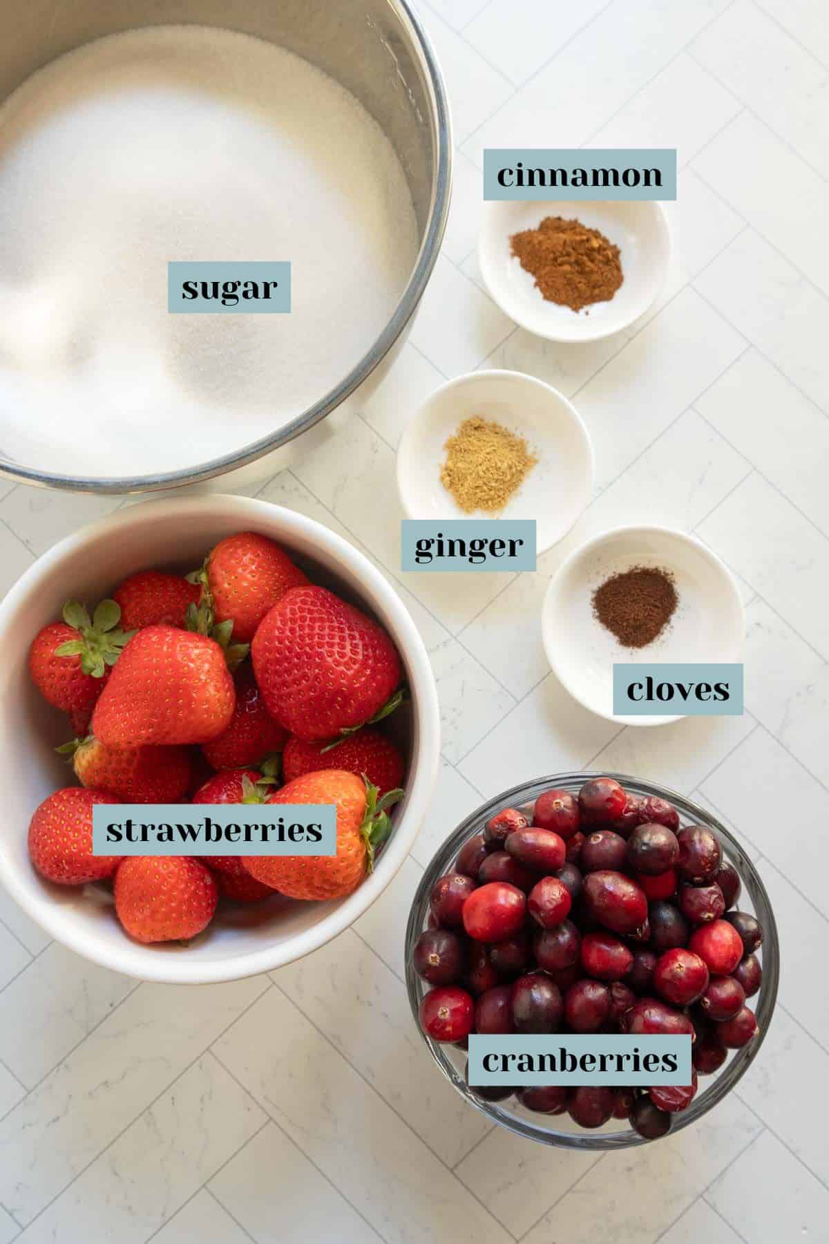 Bowls and dishes on a white surface containing sugar, cinnamon, ginger, cloves, whole strawberries, and cranberries, each labeled with text.