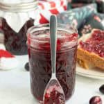 A jar of dark red jam with a spoon resting in front, surrounded by jars, a slice of bread with jam, and fresh cranberries on a white surface.