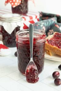 A jar of dark red jam with a spoon resting in front, surrounded by jars, a slice of bread with jam, and fresh cranberries on a white surface.