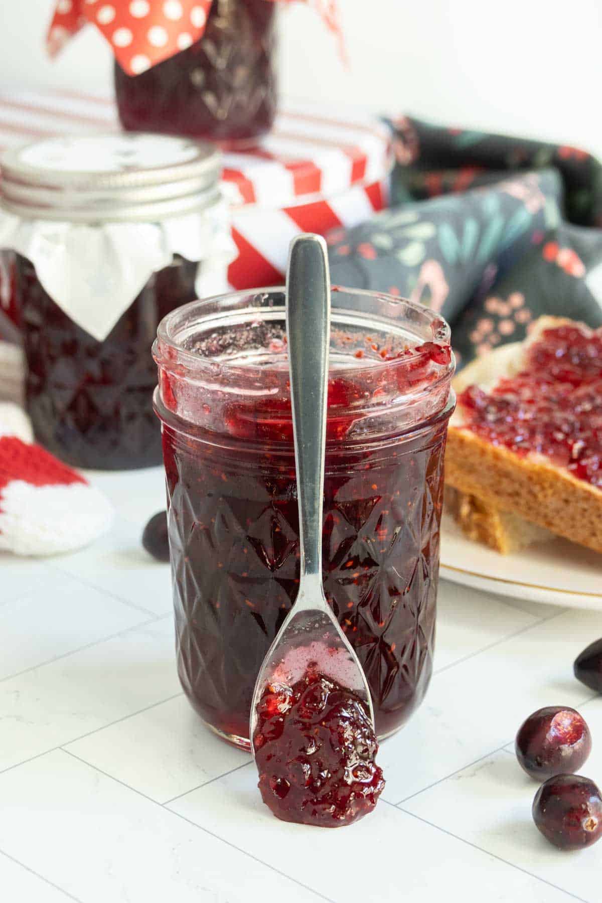 A jar of dark red jam with a spoon resting in front, surrounded by jars, a slice of bread with jam, and fresh cranberries on a white surface.