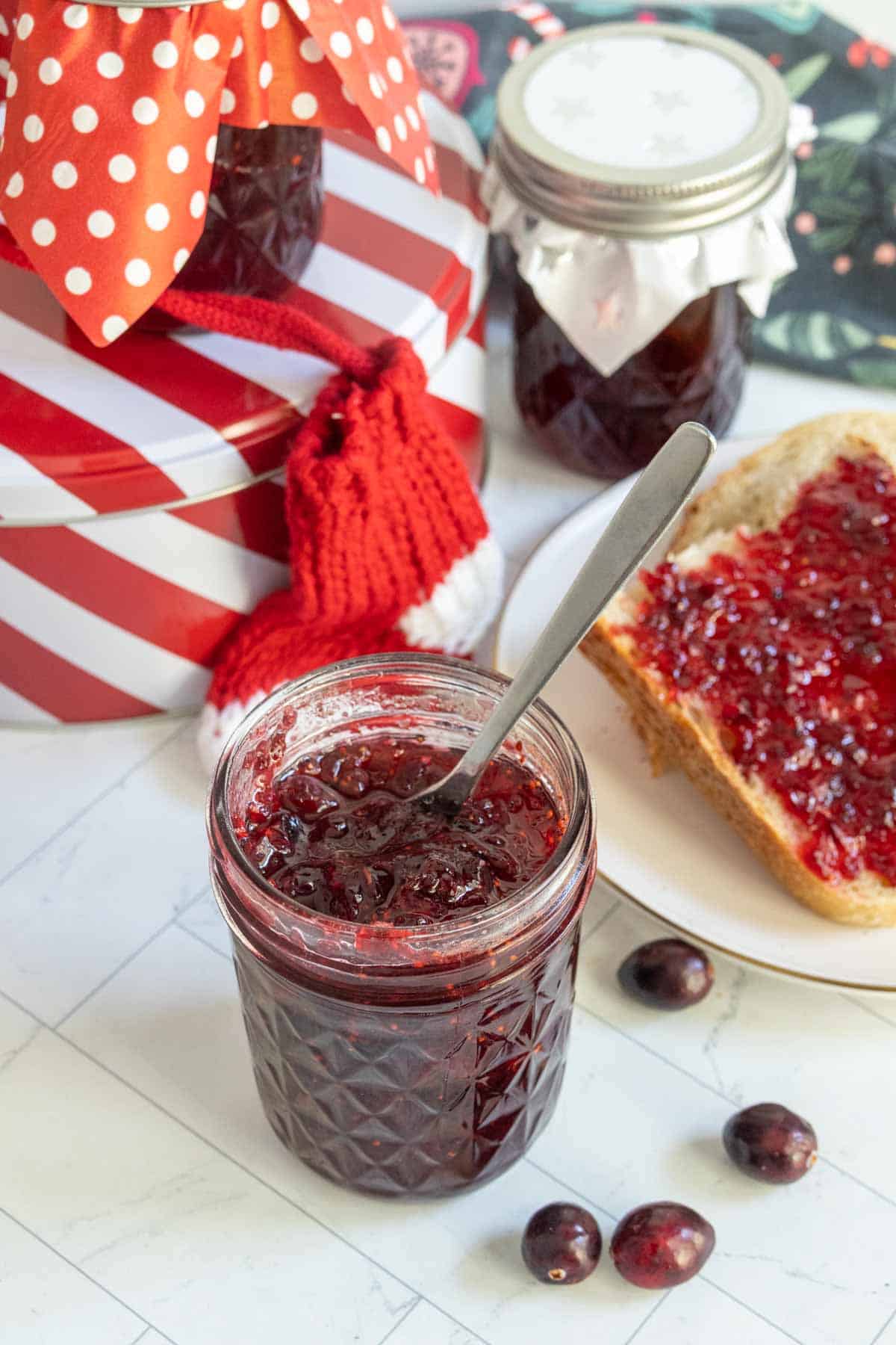 A jar of cranberry jam with a spoon inside, a slice of bread with jam on a plate, loose cranberries, and two jars with decorative lids in the background.
