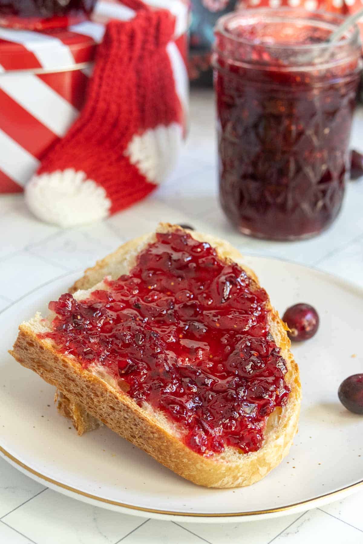 A slice of bread spread with red cranberry jam on a plate, with a jar of jam and cranberries in the background.