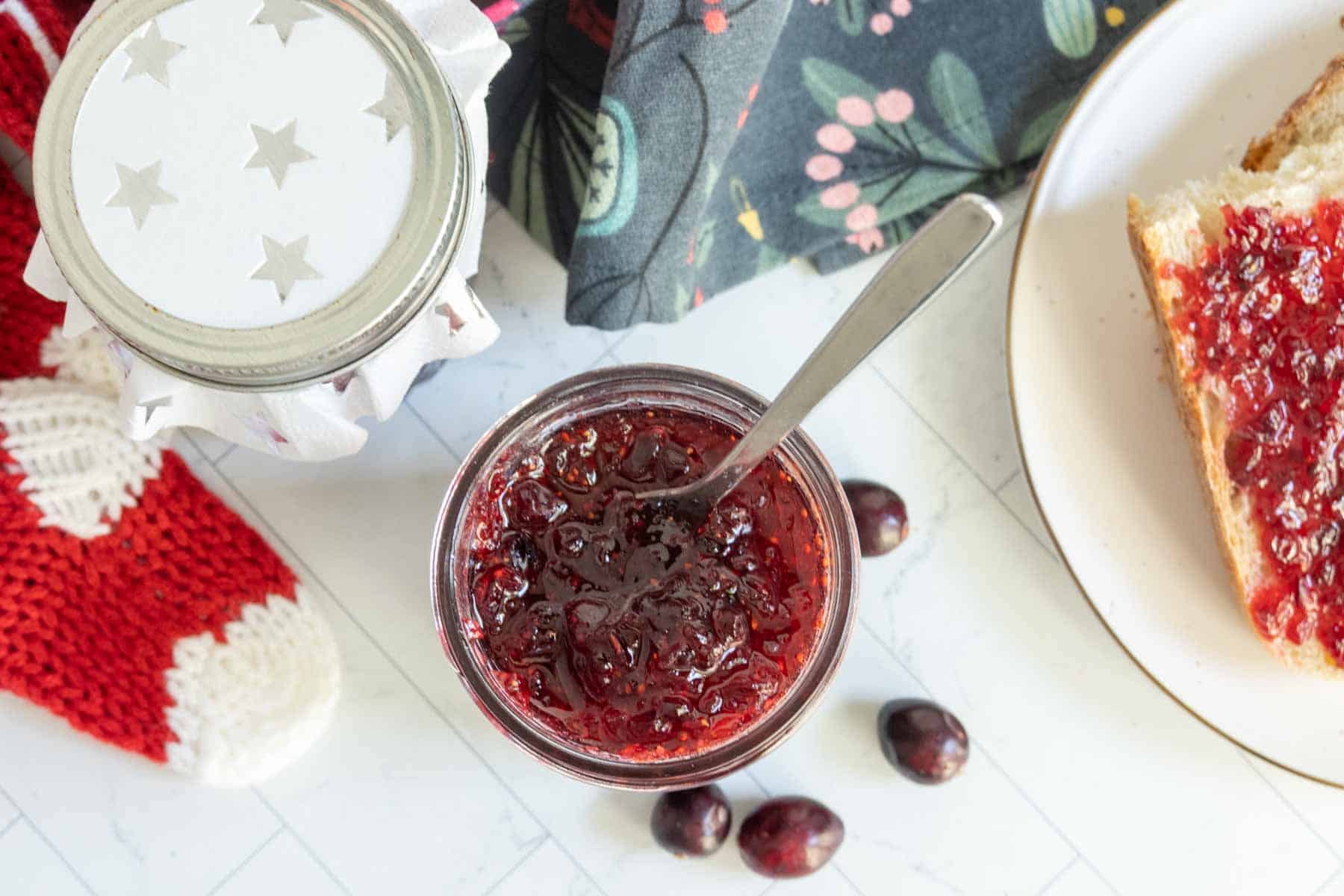 Open jar of red fruit jam with a spoon, placed next to a slice of bread with jam, cranberries, a red knitted item, and a floral-patterned cloth.