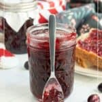A glass jar of Christmas jam with a spoon, surrounded by another jar, toast with jam, and holiday decor in the background.