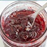 An open jar of dark red Christmas jam with visible fruit pieces, a metal spoon inside, and a festive label on the lid.