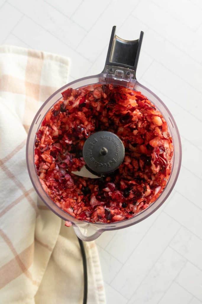 A food processor filled with finely chopped cranberries and apples sits on a light-colored cloth on a white countertop.