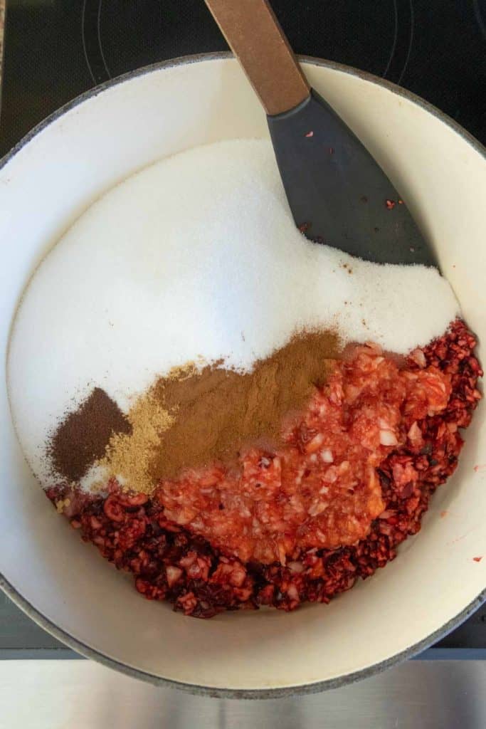 Chopped cranberries, sugar, spices, and minced onions are combined in a white pot with a black spatula resting inside.