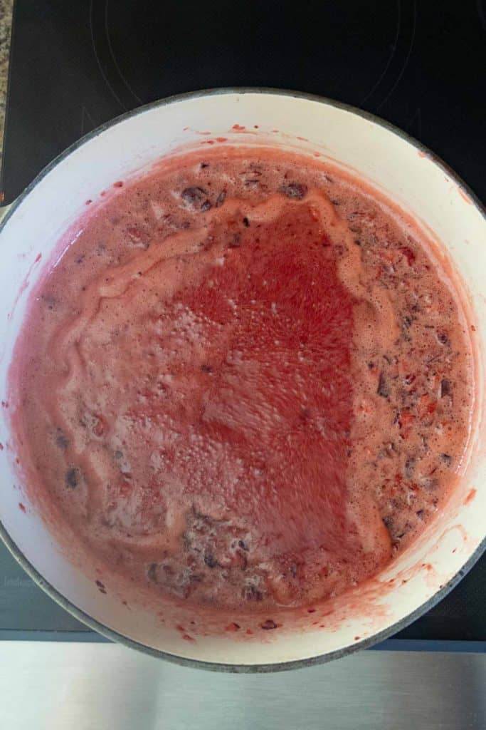A pot filled with bubbling red fruit mixture, likely simmering on a stovetop, showing both foam and liquid areas.