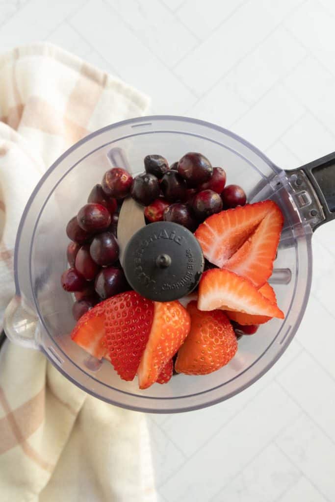 A food processor bowl contains whole cranberries and sliced strawberries, placed on a light surface next to a beige checkered cloth.