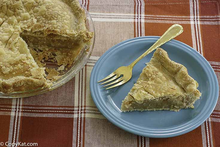 A slice of pie is served on a blue plate with a gold fork, next to a glass pie dish with the remaining pie, all placed on a red plaid tablecloth.