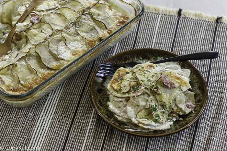 A baked dish of scalloped potatoes in a glass casserole, with a serving on a plate beside it, garnished with herbs and accompanied by a fork.