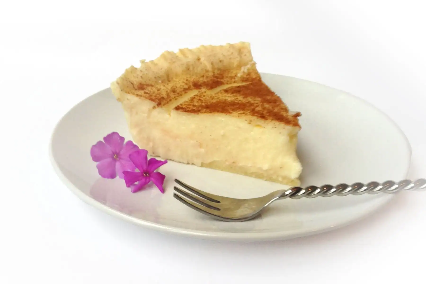 A slice of creamy pie with a dusting of cinnamon on a white plate, garnished with two small purple flowers and a fork.