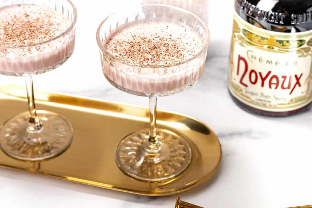 Two coupe glasses filled with a creamy, light pink cocktail topped with cocoa powder sit on a gold tray; a bottle labeled "Noyaux" is nearby.