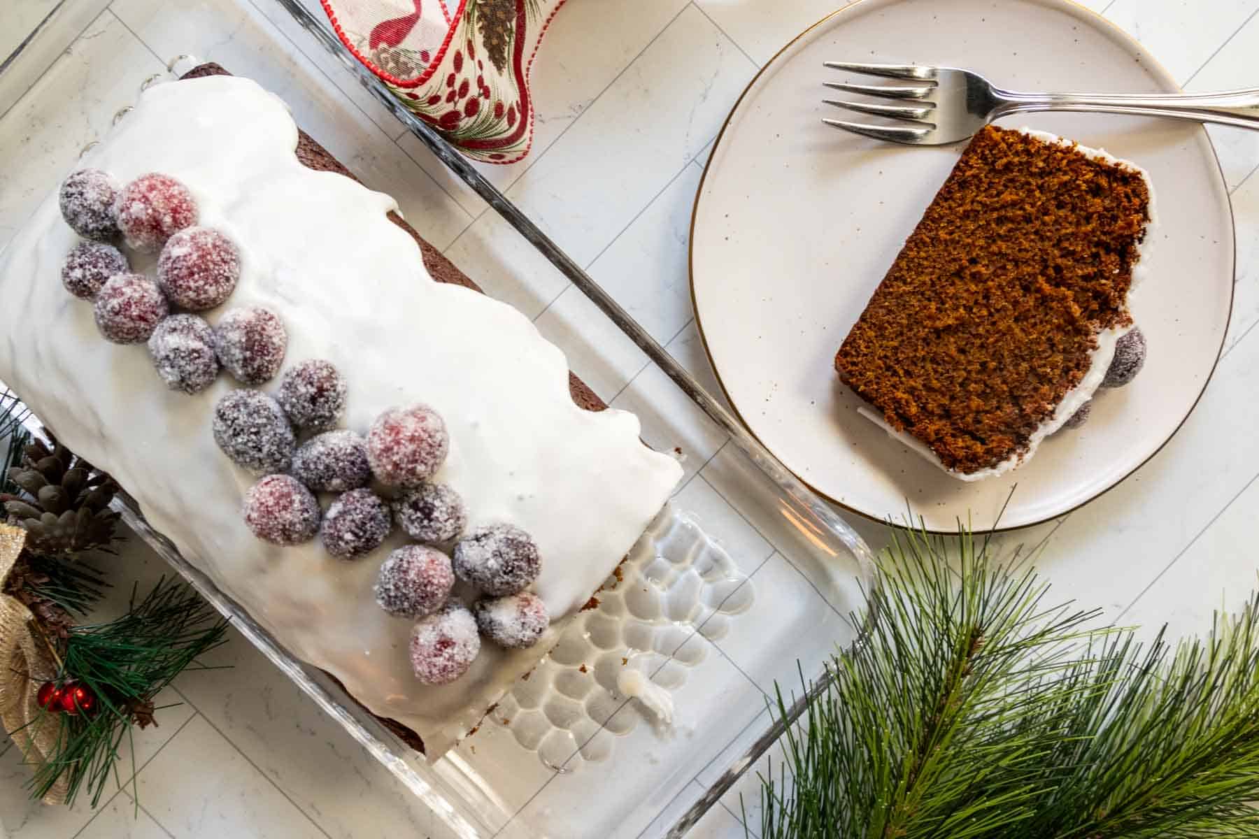A loaf cake with white icing and sugared cranberries on top sits on a glass tray; a slice is served on a plate with a fork beside it. Pine branches and holiday decor are nearby.