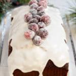 A loaf cake with white icing, topped with sugared cranberries, sits on a glass tray with holiday decorations in the background.