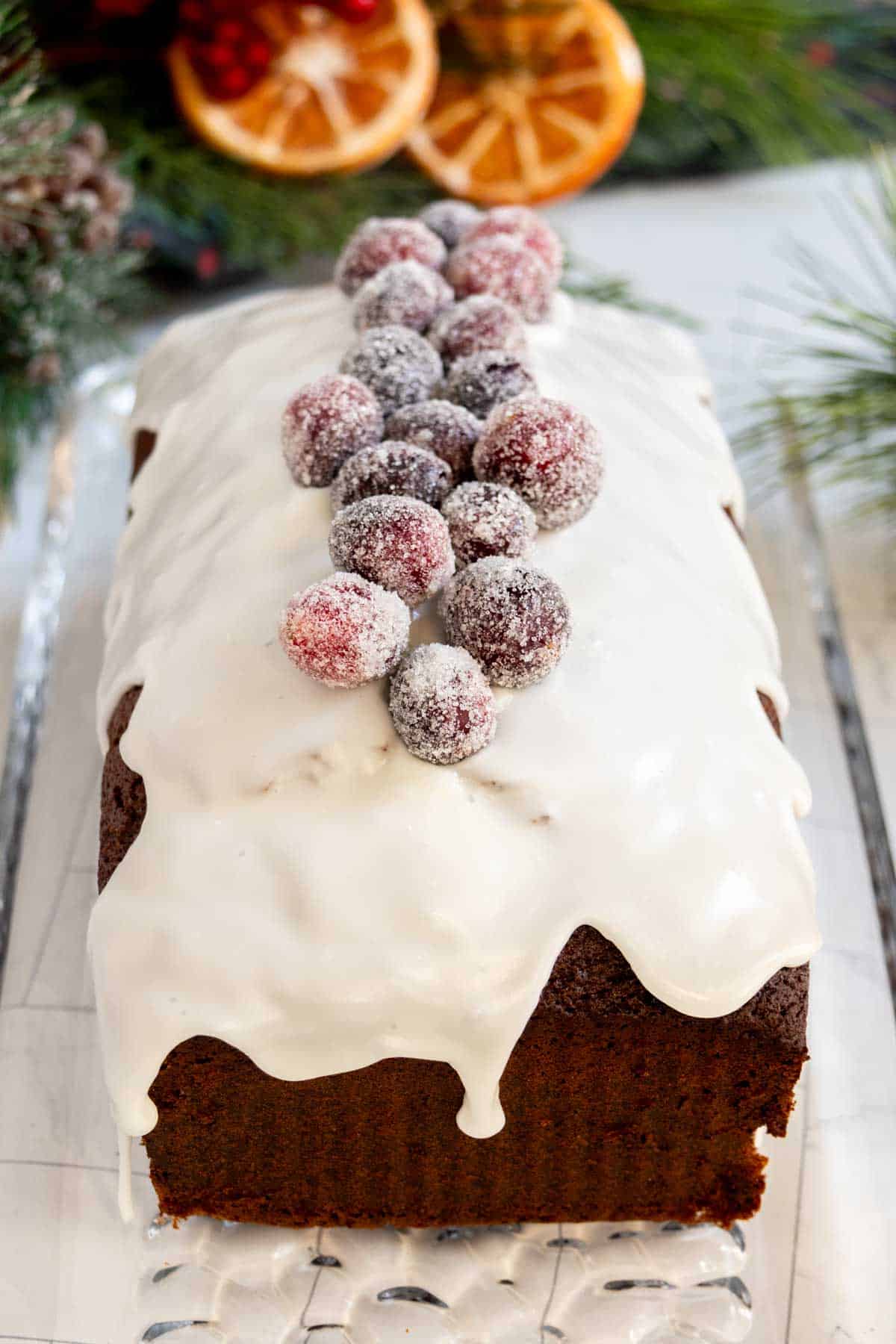 A loaf cake with white icing, topped with sugared cranberries, sits on a glass tray with holiday decorations in the background.
