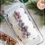 A loaf of gingerbread cake topped with white icing and sugared cranberries sits on a glass tray, surrounded by holiday decorations and greenery.