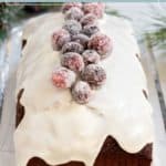 A gingerbread loaf with white icing, topped with sugared cranberries, sits on a glass tray.