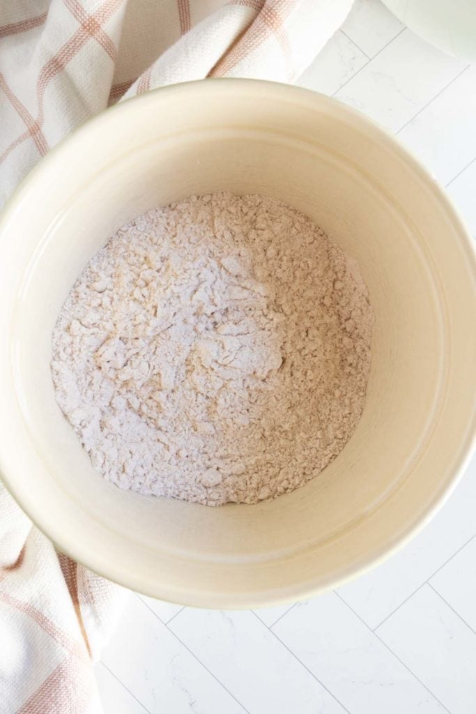A beige mixing bowl with flour and dry ingredients sits on a white surface next to a beige and pink checkered cloth.