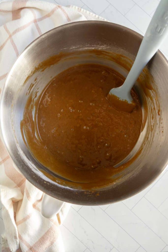 A mixing bowl filled with brown batter and a spatula resting inside, placed on a white surface with a checkered cloth beside it.