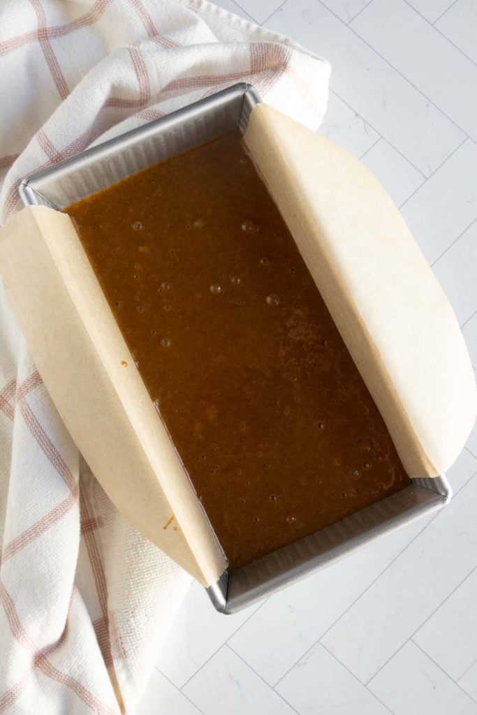A rectangular baking pan lined with parchment paper is filled with brown batter, sitting on a white towel with red stripes on a light tile surface.