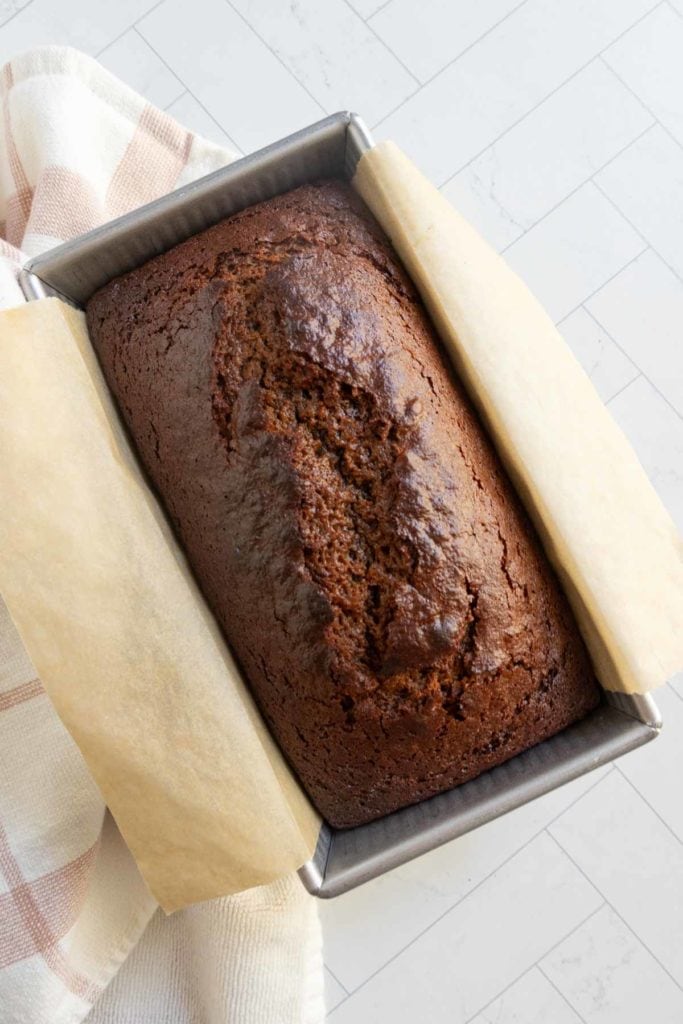 A loaf of baked banana bread sits in a parchment-lined metal pan on a light-colored tiled surface.