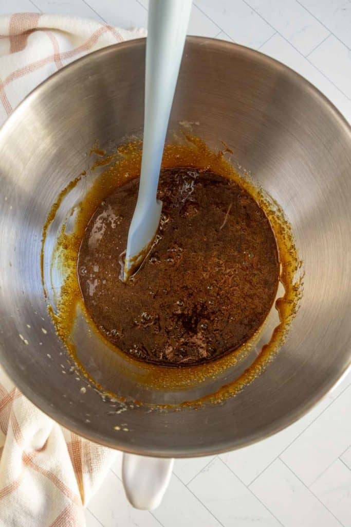 A mixing bowl containing a brown liquid mixture being stirred with a silicone spatula, placed on a white countertop with a striped towel nearby.