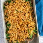A baking dish filled with green bean casserole topped with crispy fried onions, with a wooden spoon resting inside and a blue cloth nearby.