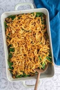 A baking dish filled with green bean casserole topped with crispy fried onions, with a wooden spoon resting inside and a blue cloth nearby.