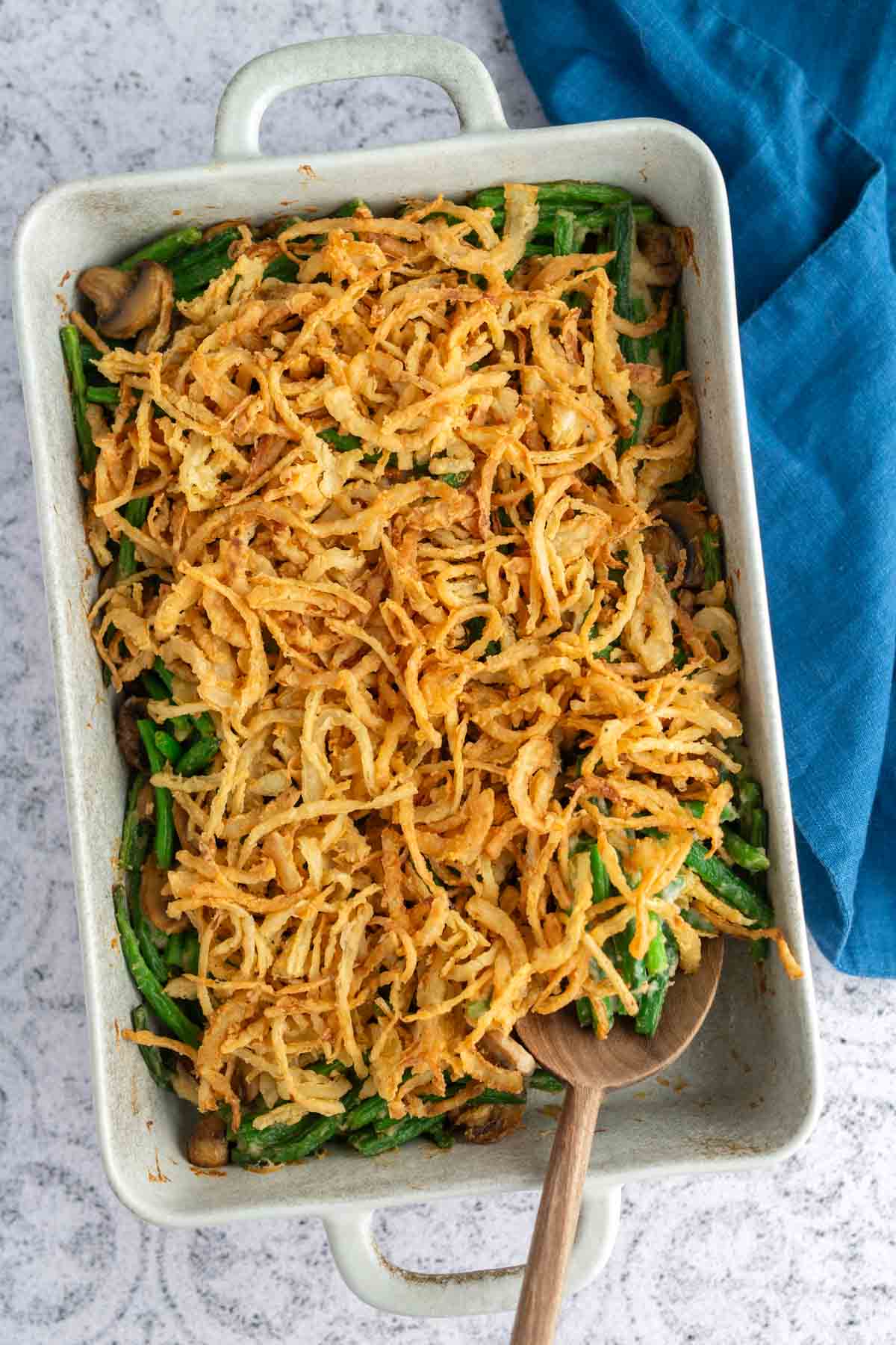 A baking dish filled with green bean casserole topped with crispy fried onions, with a wooden spoon resting inside and a blue cloth nearby.