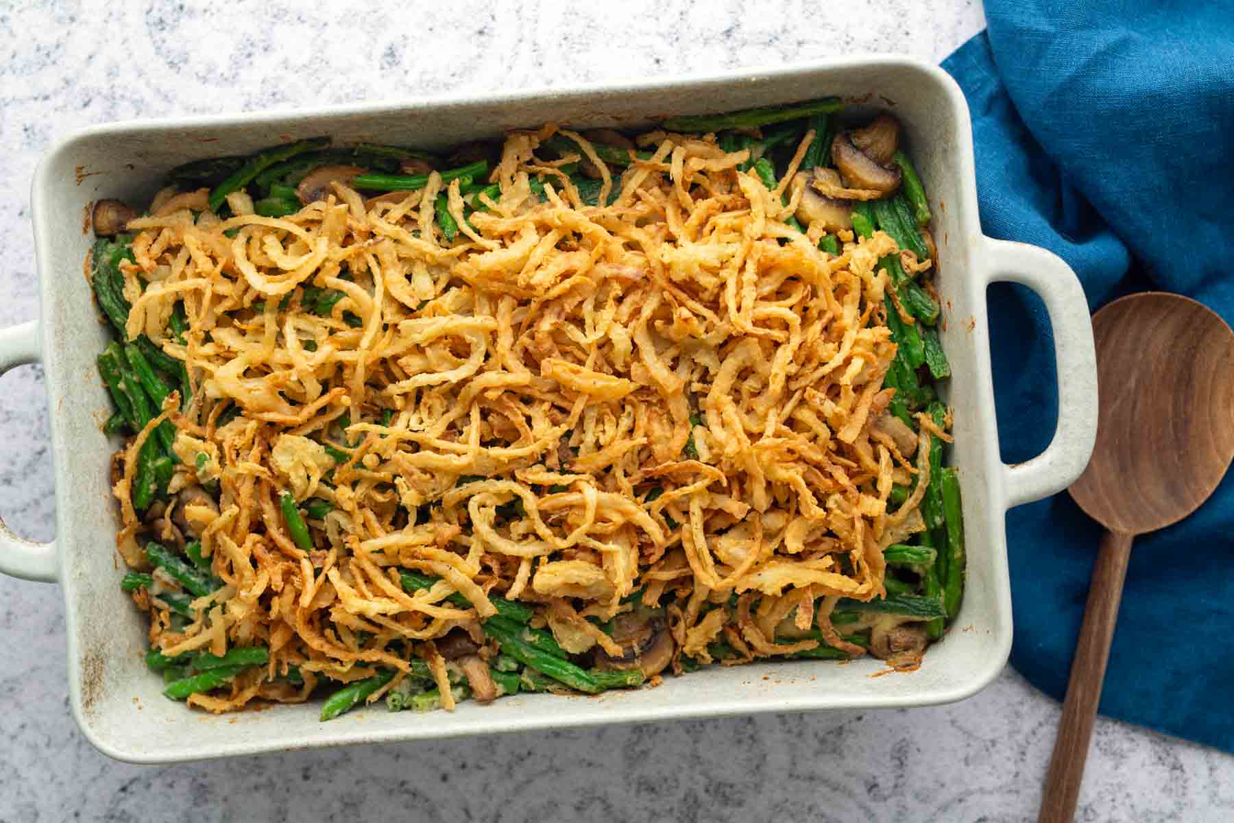 A casserole dish filled with green bean casserole topped with crispy fried onions, placed on a light surface next to a blue napkin and a wooden spoon.