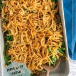 A casserole dish filled with green bean casserole topped with crispy fried onions, with a wooden serving spoon and a label stating "made with fresh green beans.
