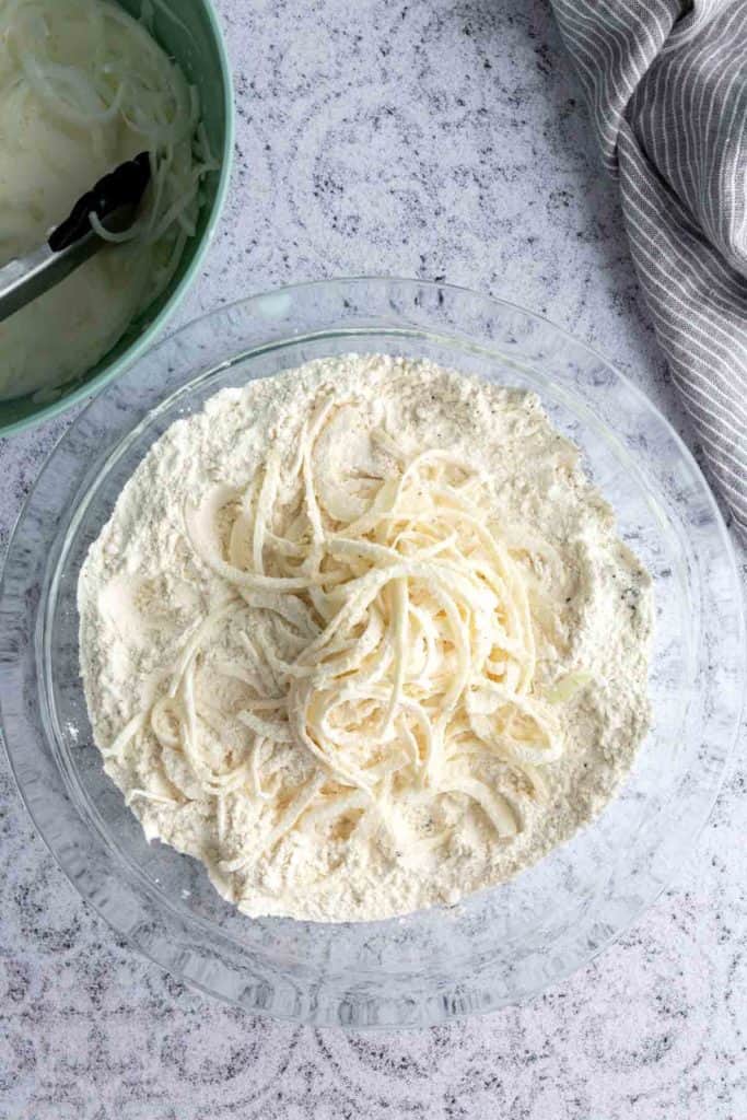 A bowl of flour with sliced onions on top, next to a green bowl containing sliced onions and liquid, and a gray striped cloth on the side.