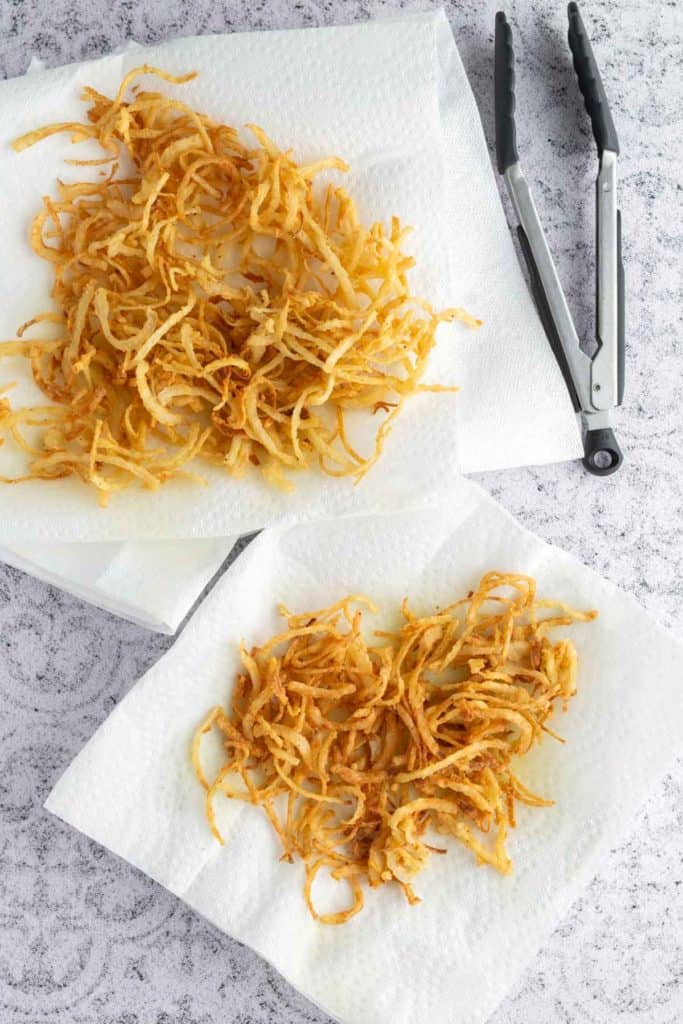 Two piles of crispy fried onion strings are placed on paper towels, with metal tongs nearby on a light gray textured surface.
