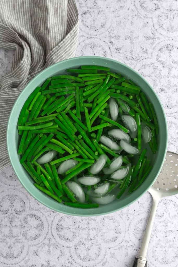 A bowl of green beans and ice cubes in water sits on a patterned surface, with a gray striped towel and a slotted spoon nearby.
