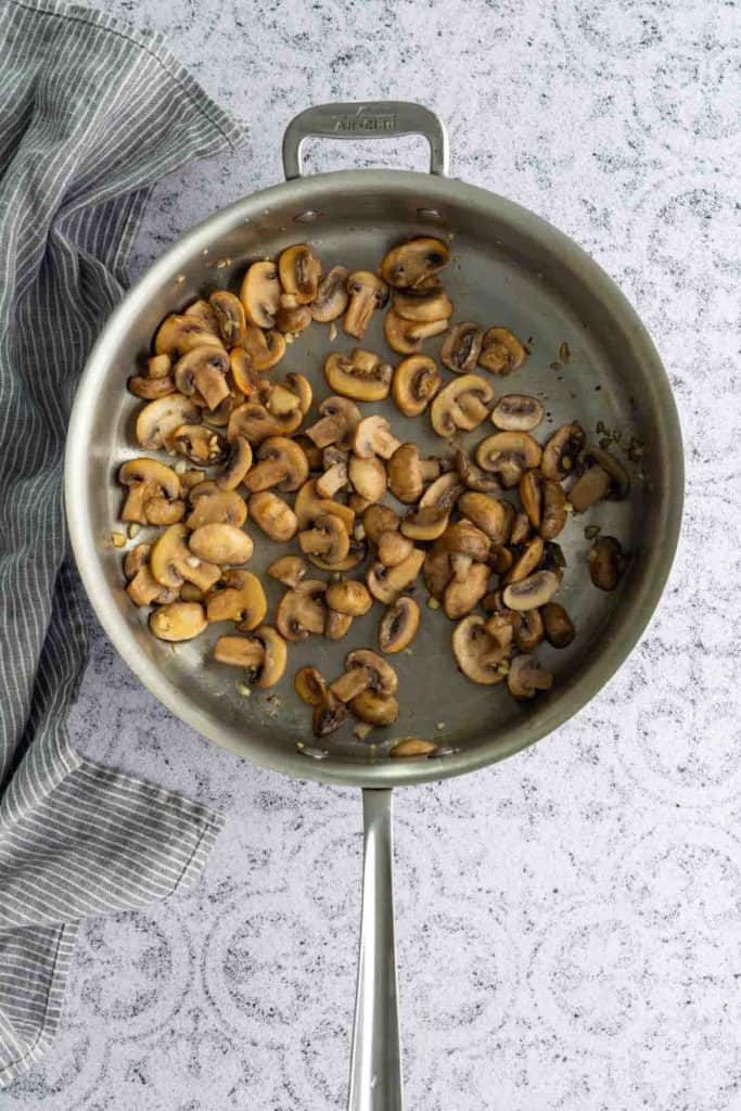 A stainless steel pan with sautéed sliced mushrooms sits on a light textured surface next to a gray and white striped cloth.