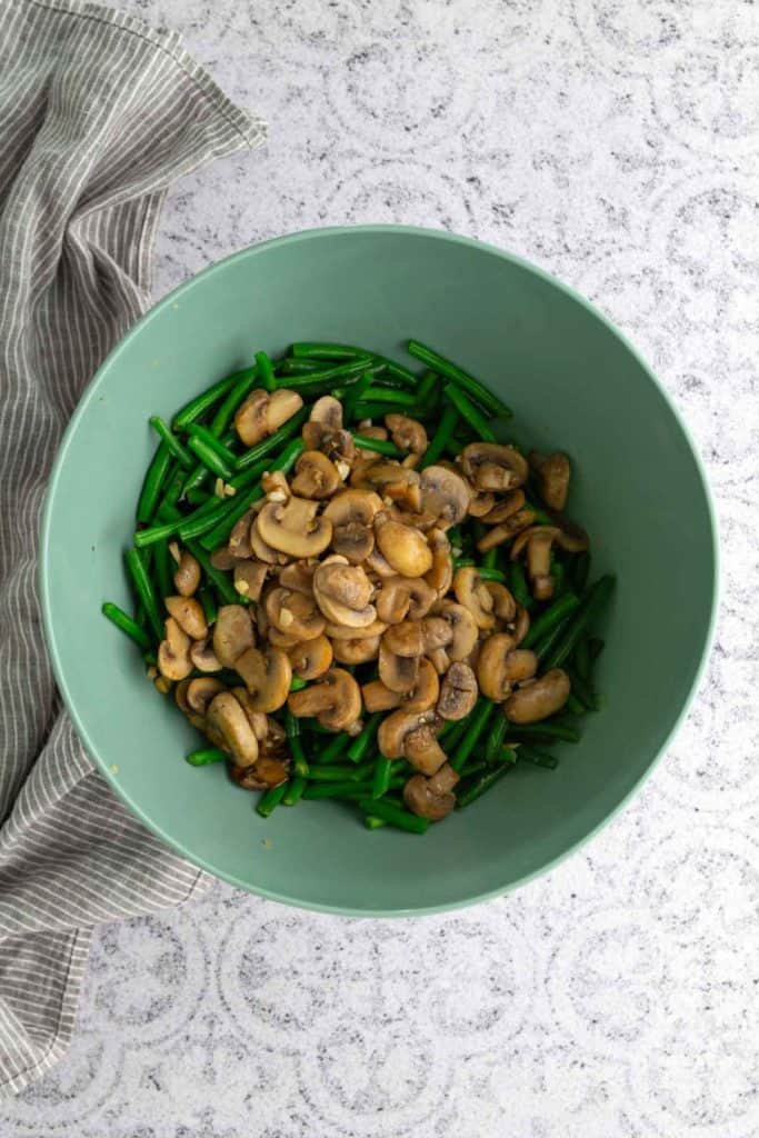 A green bowl with cooked green beans and sautéed mushrooms on a light patterned surface beside a gray striped cloth.