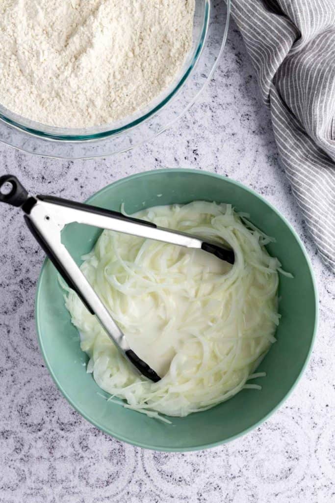 A bowl of sliced onions in liquid with metal tongs beside a bowl of flour on a gray countertop.