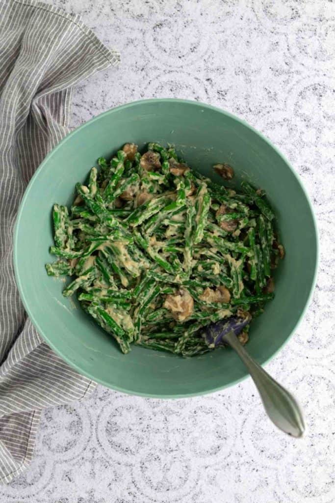 A green bowl filled with green bean casserole mixed with mushrooms and creamy sauce, next to a striped cloth on a light countertop.